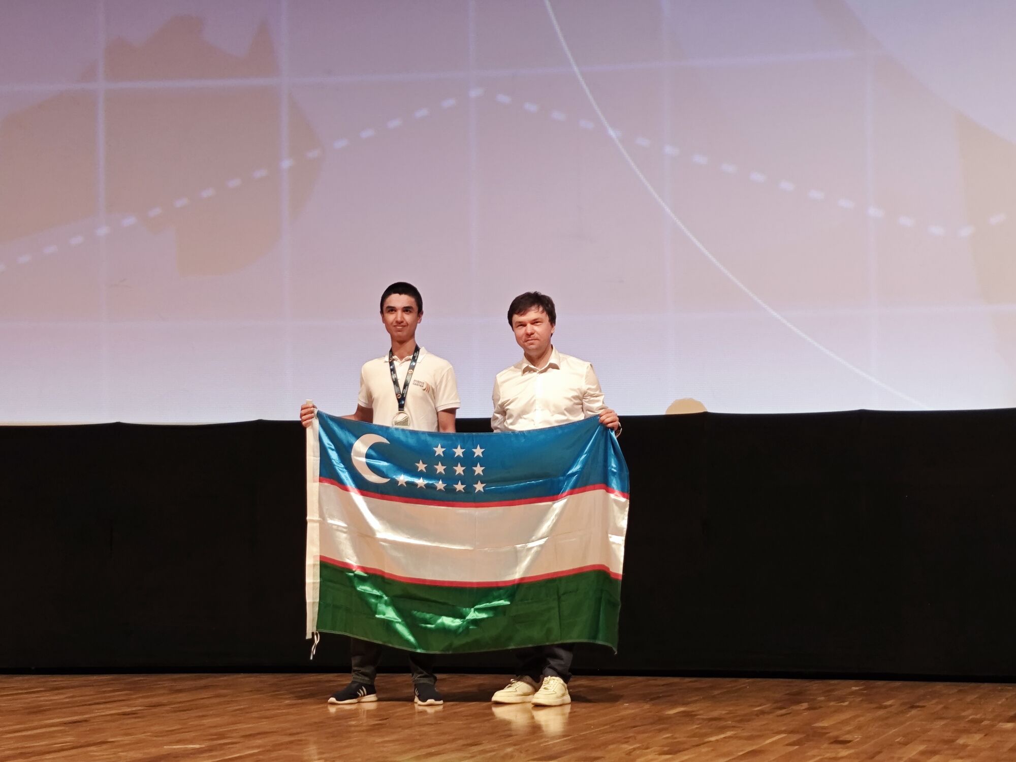 Two students on stage holding the Uzbek flag at IEO 2025